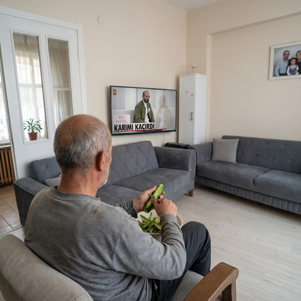 Salonda koltuğa oturmuş, salatalık soyarken televizyonda gündüz kuşağı programı izleyen bir adam
